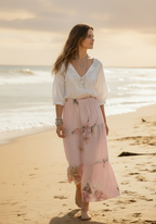 Woman in a white blouse and pink skirt standing on a beach with ocean in the background