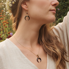 Woman wearing a gold necklace and black crescent moon earrings with a blurred natural background