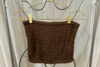 A brown smocked crop top or tube top hanging from a gold hanger with two gold clips against a light gray curtain backdrop.