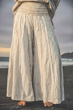Person wearing light-colored wide-leg pants standing on a beach with ocean and sky in the background