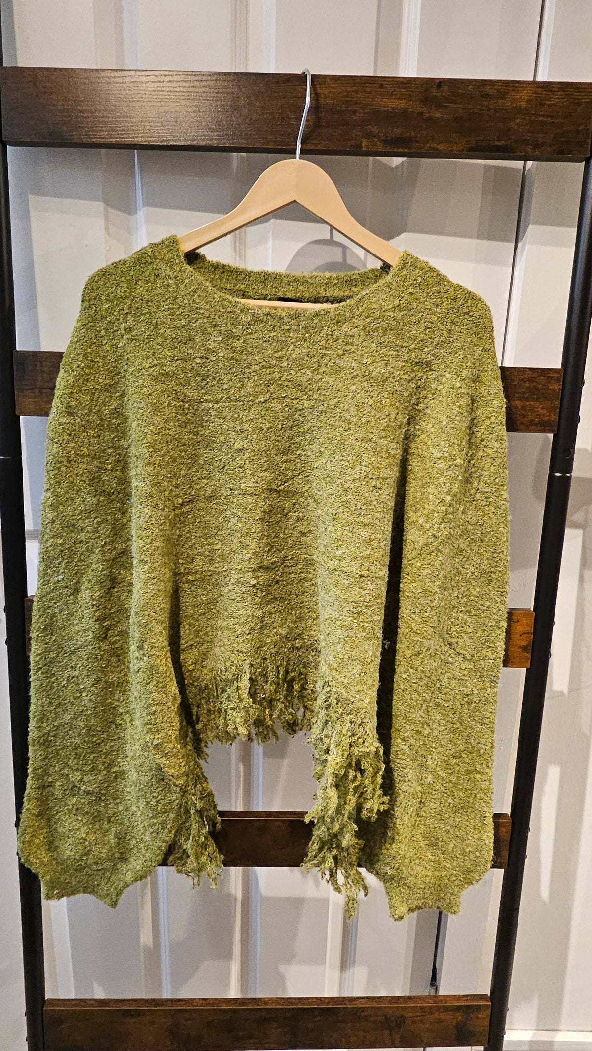 A textured, fuzzy knit sweater in a muted olive green color, hanging on a wooden hanger on a dark wooden clothing rack.