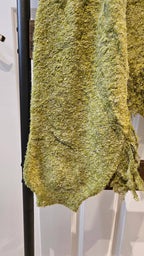 A close-up of a chunky, fuzzy knit fabric in a mottled olive green and yellow colorway, draped over a black clothing rack.