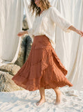 A woman standing on white sand wearing a flowing terracotta-brown tiered skirt and a loose white blouse, with draped fabric and rocks in the background.