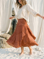 A woman standing on white sand wearing a flowing terracotta-brown tiered skirt and a loose white blouse, with draped fabric and rocks in the background.