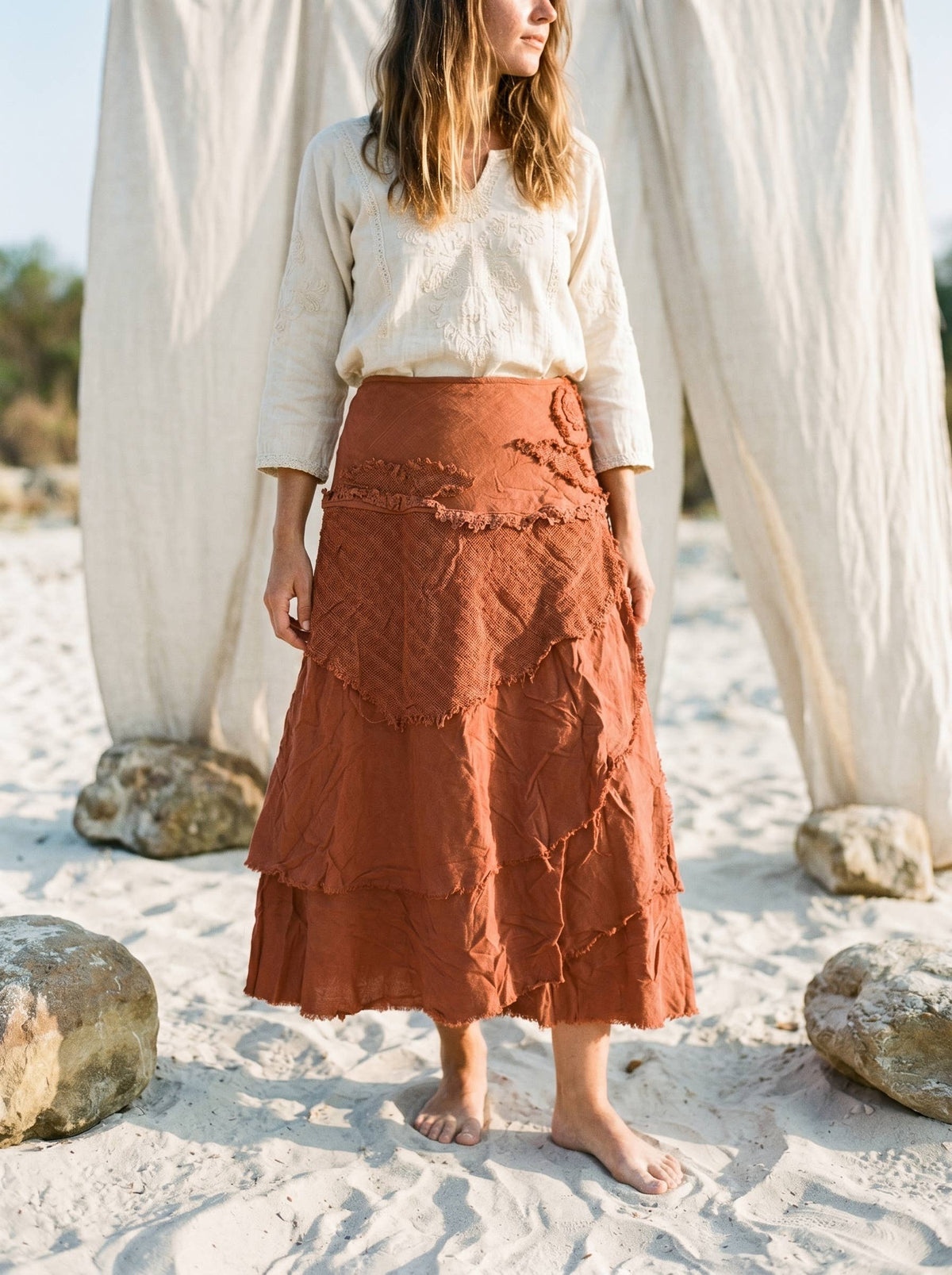 A woman standing barefoot on a sandy beach in front of draped white fabric and rocks, wearing a textured rust-orange tiered skirt with a cream long-sleeve top.
