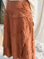 A close-up of a rust-colored, textured skirt with layered, frayed edges and floral applique details, paired with a white lace top.