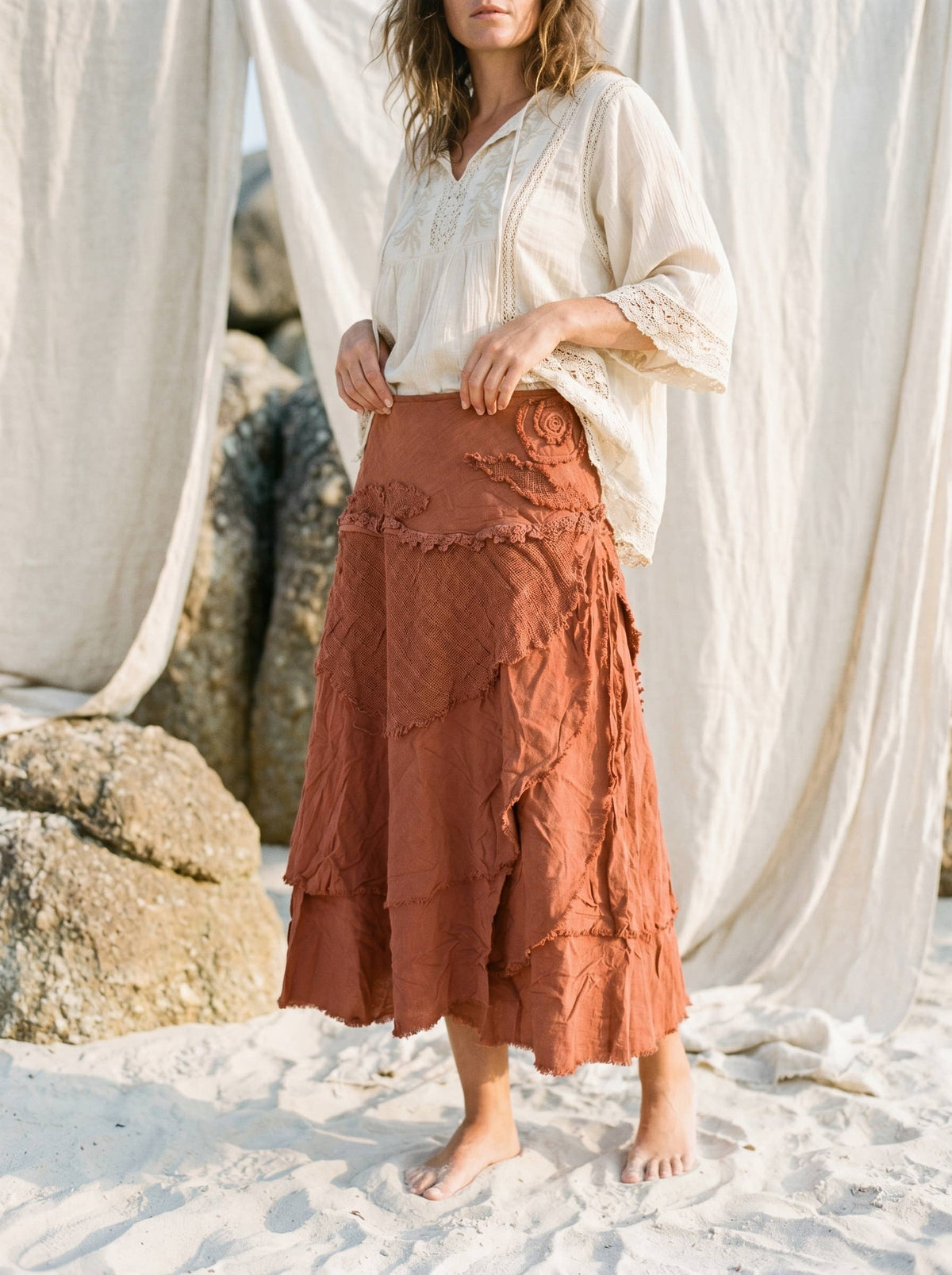 A woman standing on white sand in front of draped white fabric and rocks, wearing a loose white blouse and a rust-orange tiered skirt with textured ruffle details.
