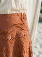 Close-up of a textured terracotta orange skirt with layered lace appliques and ruffled trim, paired with a white lace-trim top, set against a soft, warm neutral background.