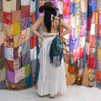 Woman in a white outfit with a brown hat and blue fringed bag standing in front of colorful fabric walls.