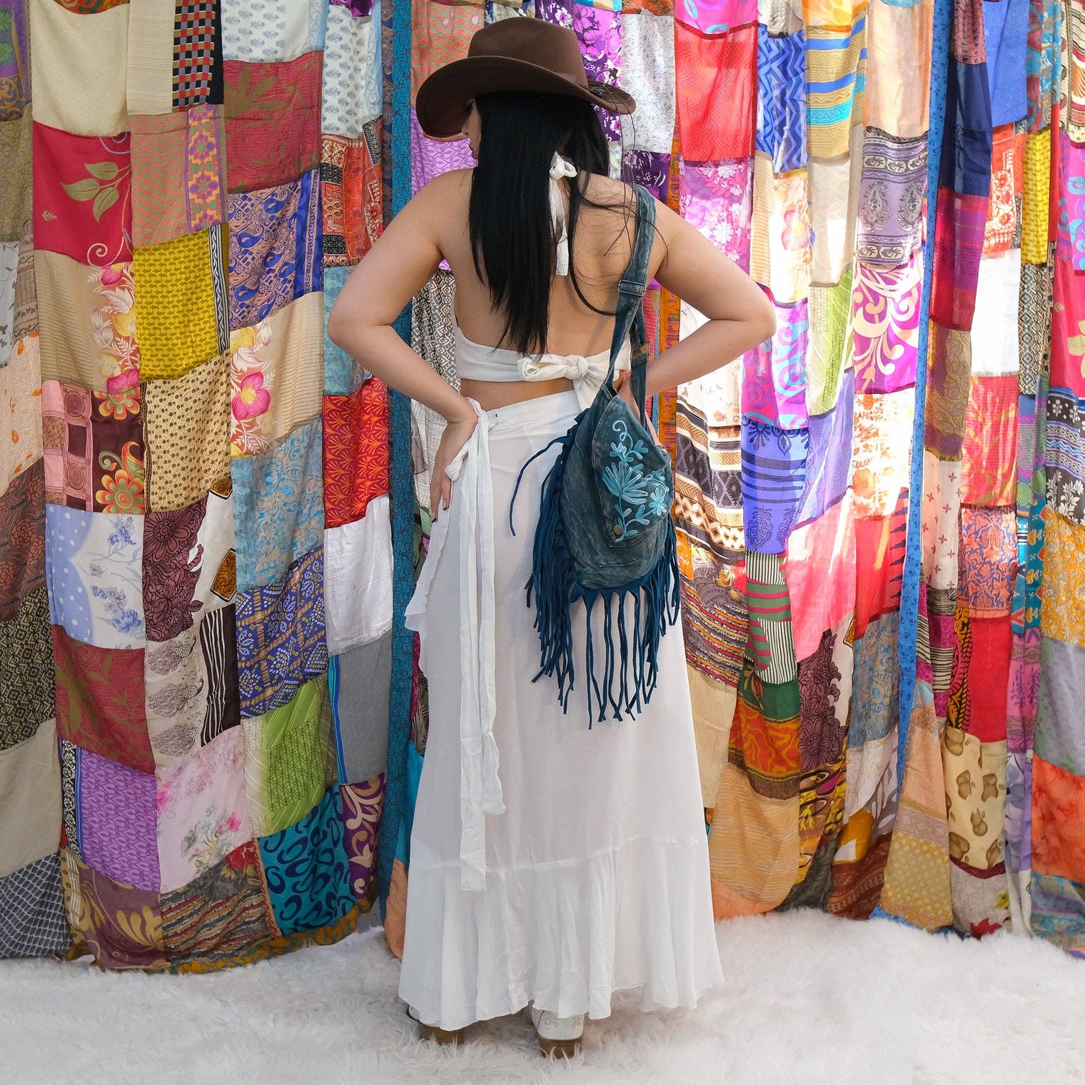 Woman in a white outfit with a brown hat and blue fringed bag standing in front of colorful fabric walls.