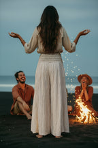 Woman in a long dress standing with arms outstretched over a campfire on a beach with two people sitting nearby.
