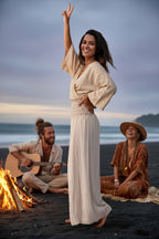 Woman in a flowing dress standing on a beach with a campfire and two people sitting in the background.