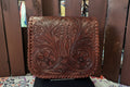 A brown leather clutch or wallet with intricate floral tooling and whipstitched edges, displayed on a stand in front of a wooden slat background.