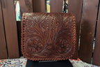 A brown leather clutch or wallet with intricate floral tooling and whipstitched edges, displayed on a stand in front of a wooden slat background.