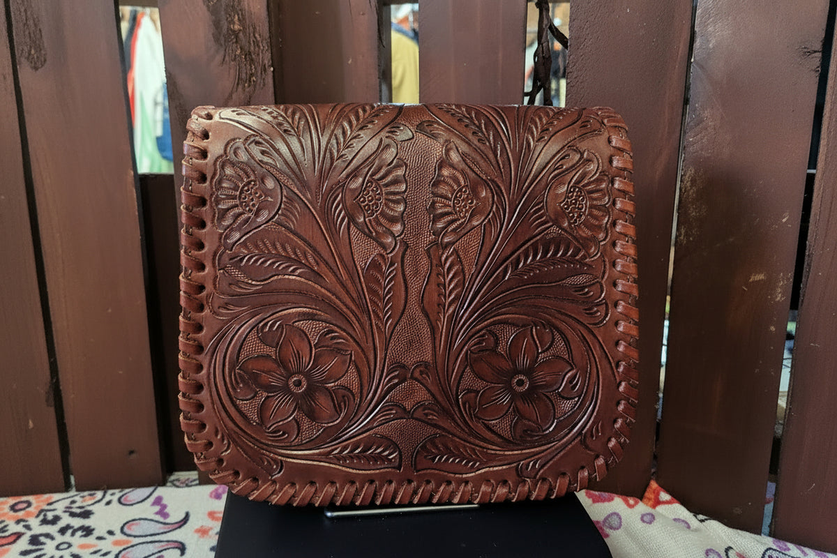 A brown leather clutch or wallet with intricate floral tooling and whipstitched edges, displayed on a stand in front of a wooden slat background.