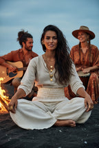 Woman sitting cross-legged in front of a fire with two people playing instruments in the background.